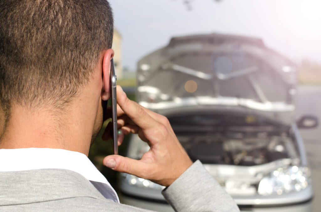 Homem parado em frente a um veículo quebrado, falando no celular